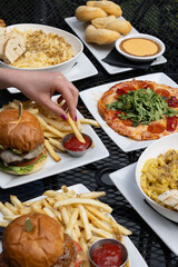 Hand dipping french fry at outdoor table with burgers, fries, mac and cheese, pizza, and pretzels