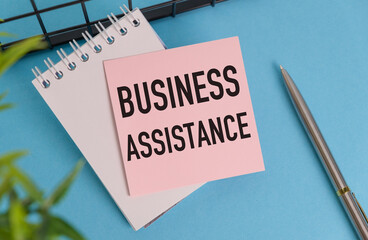 A pink sticky note with the word Business Assistance written on it is sitting on top of a spiral bound notebook.