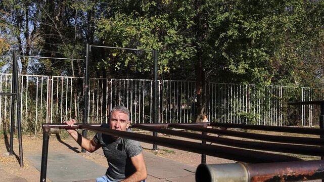 54-Year-Old Fit Man Doing Crunches on Parallel Bars - Wide Angle - Sunny Day. Man in excellent physical condition doing crunches on low parallel bars. Suitable for core strength and fitness promotion.