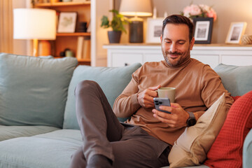 Portrait of confident businessman holding coffee and texting over cellphone while sitting on sofa at home