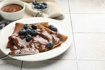 Delicious chocolate crepes with sweet paste and blueberries on light tiled table, closeup. Space for text
