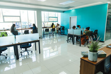 General view of a naturally lit coworking space, featuring professionals working at shared desks and high tables next to large windows.