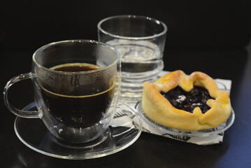 A double-walled glass cup of coffee, a glass of water, and a blueberry pastry on a small plate, all set on a black table for a simple and inviting scene