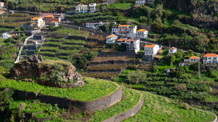A picturesque hillside village with white houses and red roofs, surrounded by terraced green fields and stone walls. The scene captures the charm of rural living in a lush landscape.