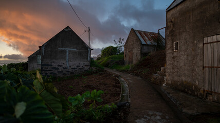 Fototapeta premium A quiet rural scene at sunset, featuring old stone farmhouses along a dirt path. The sky glows with warm hues, contrasting with the earthy tones of the buildings and surrounding greenery.