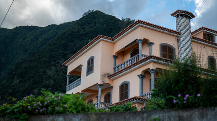 A charming peach-colored house with decorative columns and a striped chimney, set against a backdrop of lush green mountains and a partly cloudy sky, creating a serene and picturesque setting.