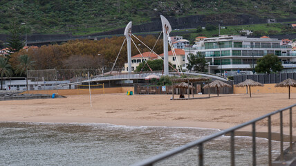 A quiet sandy beach with straw umbrellas, a modern pedestrian bridge, and nearby buildings, framed by green hills under an overcast sky.