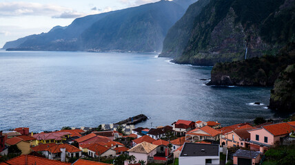 A coastal village with red-roofed houses overlooking the ocean, surrounded by dramatic cliffs and lush greenery. A tranquil blend of natural beauty and charming architecture.