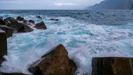Waves crashing against large rocks along a rugged coastline, with foamy water and distant cliffs under a cloudy sky. A dynamic seascape showcasing nature’s power.