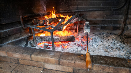 A close-up of a traditional fireplace with burning wood on a metal grate, glowing embers, and ashes. A wooden-handled tool rests nearby, evoking warmth and coziness.
