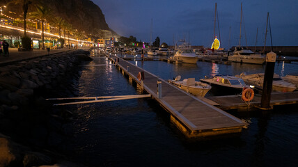 A marina at dusk with illuminated boats and a wooden dock reflecting on calm water. Coastal lights and a rocky hill create a peaceful evening ambiance.