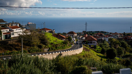 A scenic coastal village with red-roofed houses on lush green hills overlooking the ocean. A winding road and a stone wall add charm to the tranquil rural setting.