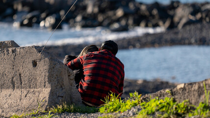 A couple sitting by the water, with one wearing a red plaid shirt, enjoying a quiet moment near a rocky shoreline. The scene captures a serene and intimate coastal atmosphere.
