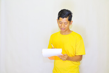 Young man in a bright yellow t-shirt carefully making notes on a rolled up piece of paper, a thoughtful expression on his face against a plain white background,