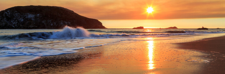 A sunset on the beach with a breaking wave and sea stacks in the distance with a reflection of the...