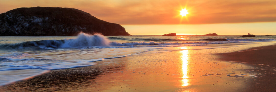 A sunset on the beach with a breaking wave and sea stacks in the distance with a reflection of the sun on the wet sand.