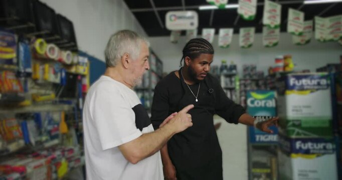 Older man and younger male worker pointing at paint cans in a hardware store aisle, discussing products, diverse teamwork in professional retail environment