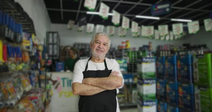 Older male store owner crossing arms confidently in hardware store aisle, representing leadership and pride in diverse retail business setting