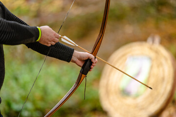 Archer's hand with wooden bow and arrow before shooting with an outdoor target in the background.