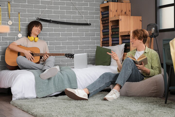 Male student playing guitar and his friend reading book in bedroom