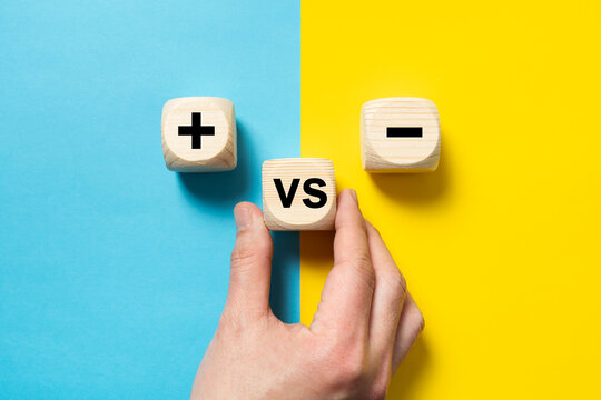 Hand placing a wood block versus between plus and minus sign on blue and yellow background, concept of opposites, pros and cons, lay flat composition
