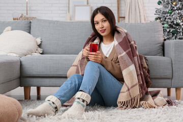 Beautiful young happy Asian woman with cup of hot mulled wine and plaid sitting on floor at home
