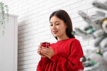 Beautiful young happy Asian woman with cup of hot mulled wine near Christmas tree at home