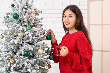 Beautiful young happy Asian woman with cup of hot mulled wine decorating Christmas tree at home