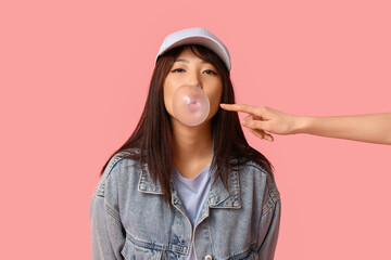 Pretty young Asian woman blowing bubble gum and female hand on pink background