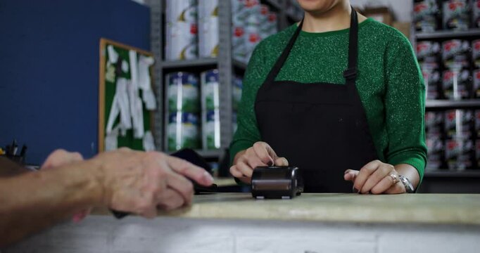 Close-up of an older man making a payment using a card machine at a hardware store counter, while a woman assists in the transaction