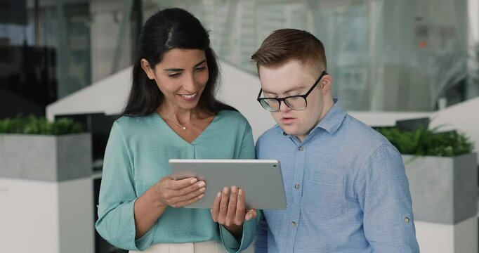 Young skilled professional woman explaining task or new platform to young man with Down syndrome, collaborating using digital tablet together during workday in office, engaging in learning or teamwork