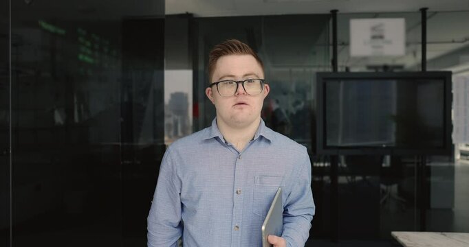 Portrait of young smiling man with congenital disability Down syndrome, dressed in casual shirt wear eyeglasses posing for camera standing alone in modern workspace with digital tablet. Inclusivity