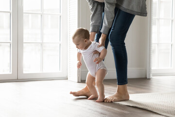Making first steps. Cute little infant baby boy or girl toddle barefoot at light living room on warm wooden floor with heating learn to walk feeling help support of caring mom nanny hands. Copy space