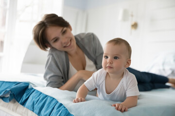 Obraz premium Child development and safety. Smiling young mom nanny lying on big comfy bed watching cute infant babe learning to crawl. Focus on baby kid standing on all four looking forward with cognitive interest