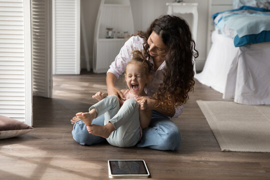 Tickling giggling having fun. Affectionate millennial mum nanny play active game messing kidding with laughing small kid girl preschooler on floor with heating at bedroom distracted from tablet screen - Powered by Adobe