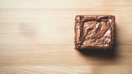 A single chocolate brownie placed on a light wooden surface with negative space around.