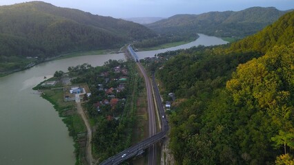 Aerial drone view of the Serayu River, the highway and railroad tracks on the side, with a background of hills, green pine forests and bright clouds in the afternoon