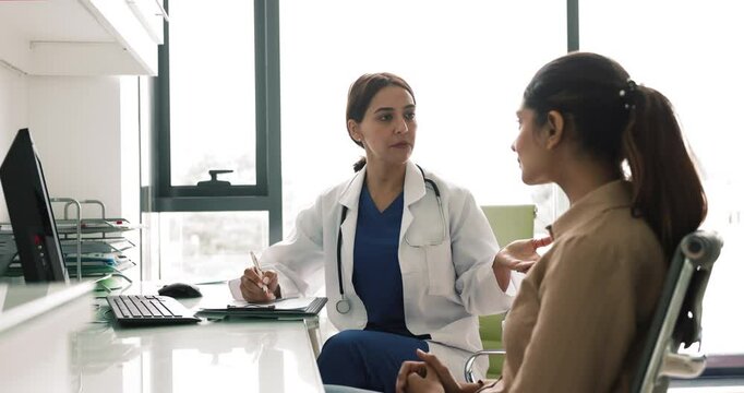 Serious Middle Eastern female doctor talking to patient woman about healthcare complaints, sitting at workplace table, giving consultation in hospital office, writing notes, keeping medical records