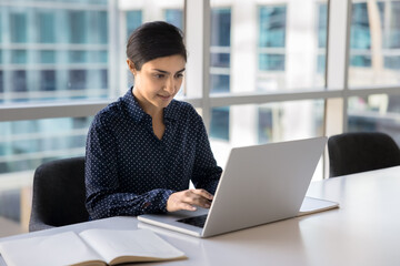 Positive busy young Indian businesswoman typing on laptop in office, sitting at workplace table with large window behind, working on online project, using technology for job communication