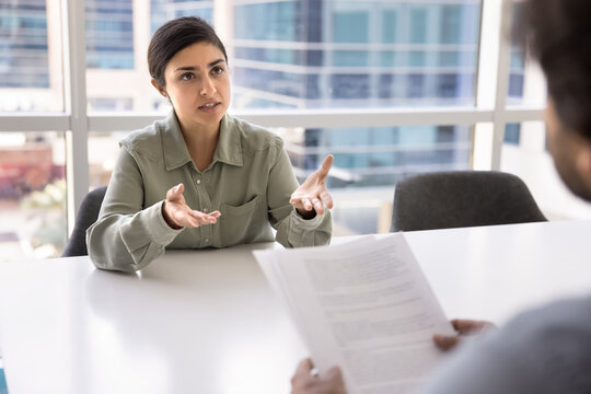 Serious beautiful young businesswoman talking to male business colleague at meeting table, speaking at job interview, consulting legal advisor, lawyer holding paper document