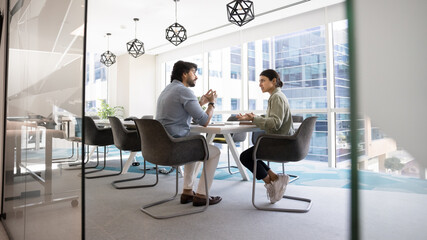 Two male and female coworkers talking at large conference table in contemporary office interior, meeting for teamwork, creative planning. Wide banner shot through glass doorway