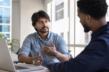 Confident Indian business project leader man talking to African partner workplace table, explaining...