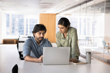 Two Indian business partners meeting at workplace table, talking at laptop, looking at screen, speaking, discussing online startup content, Internet marketing strategy in co-working office