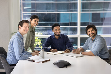 Happy multiethnic business colleagues sitting at large table in conference meeting room, looking away together, smiling, laughing, watching presentation, glad to see someone, enjoying cooperation