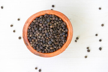A clay cup filled with aromatic black peppercorns (Black Pepper) on a light wooden surface. Top view.