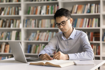 Engaged in learning. Serious motivated young male student wearing glasses study literature at library desk do assignment write in copybook take notes use notebook to get information online. Copy space