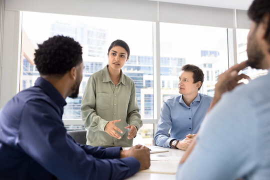 Inspired young Indian manager woman speaking to colleagues, telling creative ideas for business plan. Multiethnic team of coworkers meeting in conference room for teamwork, brainstorming