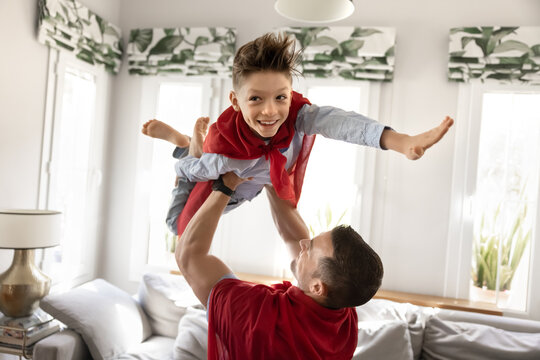 Strong caring father lifting cheerful son wearing superhero cape overhead. Happy playful boy keeping balance, making flying superhero hand gesture, smiling, laughing