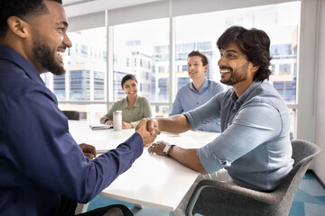 Happy confident diverse business colleagues shaking hands at conference meeting, smiling, enjoying cooperation, partnership. Indian boss thanking African manager for good job, giving handshake