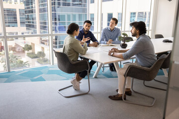 Diverse team of young male colleagues listening to speaking Indian business leader at meeting table in collaboration room interior, talking in contemporary workspace. Candid shot with copy space